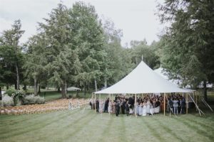 A large white tent on a grassy lawn shelters a gathering of people dressed formally, with rows of empty wooden chairs and tall green trees surrounding the outdoor wedding ceremony, perfect for those seeking customizable packages.
