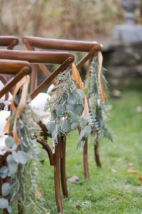 Wooden chairs arranged outdoors, each decorated with green eucalyptus leaves and tied with peach-colored ribbons, set on a grassy area for an event or wedding ceremony.