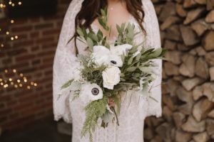 A bride in a lace dress holds a large bouquet of white flowers and greenery. Stacked firewood and exposed brick are visible in the background.