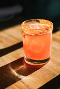 A glass of pink cocktail with ice cubes, served on a wooden surface. The rim of the glass is coated with chili or spice seasoning, making it a vibrant drink perfect for any menu, highlighted beautifully by the sunlight.