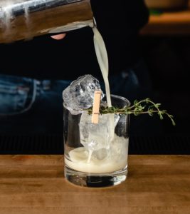 A creamy cocktail is being poured from a shaker into a glass with large ice cubes, a small clothespin, and a sprig of fresh thyme, ready to delight as the newest addition to the menu on the wooden bar counter.