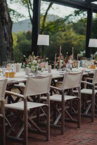 An elegant outdoor dining table set with white chairs, floral centerpieces, tall candles, and glassware, surrounded by greenery and trees in a natural, scenic setting.
