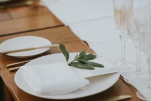 A close-up of an elegant weddings table setting with a white plate, folded white napkin, small green leaf, gold cutlery, a white card, and crystal glasses on a wooden table with a white table runner.