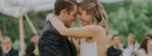 A bride and groom smile with their foreheads touching as they share a close dance outdoors, surrounded by guests seated at tables in a lush, green setting—capturing the magic included in our wedding packages.