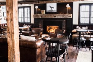 A cozy lounge with leather armchairs, wooden tables, and a lit fireplace with a rustic wooden mantel. Black chairs surround tables where guests can relax or open packages. A crow painting hangs above the fireplace, and windows let in natural light.