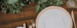 A rustic wooden table set for weddings, featuring a white plate, beige charger, white napkin, and gold utensils, complemented by a glass of water and green leafy garland as decoration.