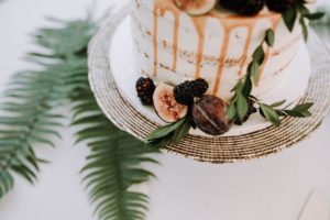 A white frosted cake with caramel drizzle, decorated with figs, blackberries, and green leaves, sits on a decorative platter—perfect for weddings. Fern leaves are arranged on the white surface underneath.