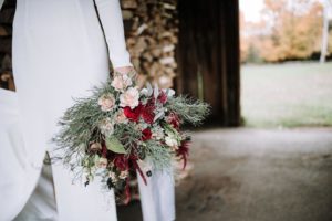 A person in a white dress holds a large, rustic bouquet of blush pink and deep red flowers with greenery, standing in a wooden structure with stacked logs and autumn trees in the background.