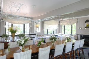 A bright dining room with a long wooden table, white chairs, hanging glass globes, and white decorative branches overhead. The table is adorned with vases of fresh flowers, gold candle holders, and charming gift packages beneath large sunlit windows.