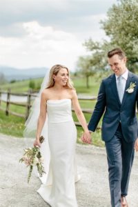 A bride in a strapless white gown and veil holds a bouquet and walks hand-in-hand with a groom in a navy suit and tie. They are smiling outdoors on a path with a wooden fence and greenery in the background.