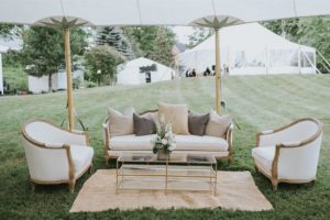 A stylish outdoor lounge setup, perfect for weddings, features a white sofa, two matching armchairs, a glass coffee table with a floral arrangement, and a tan rug on green grass beneath a canopy with large white tents in the background.