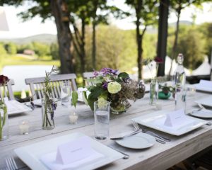 A wooden outdoor table is set with white plates, glassware, name cards, and small flower arrangements in jars. Packages are neatly placed among the decor, perfect for a garden or outdoor event surrounded by trees and greenery.