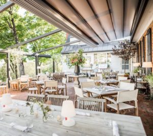 Open-air restaurant patio with white tablecloths, wooden chairs, and soft lighting. Tables set with glasses and napkins are surrounded by greenery and flowers, under a partially covered roof. Special packages available for groups on sunny days.