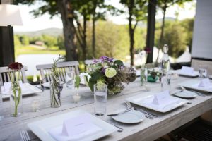 A beautifully set outdoor dining table with white plates, glasses of water, place cards, and vases of flowers. The background shows trees, greenery, and a view of a lake or pond.