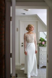 A bride stands in a white room, wearing a white wedding dress with a sheer, floral-embroidered bodice and long sleeves. She is looking to the side and smiling, with a bouquet of flowers on a dresser nearby.