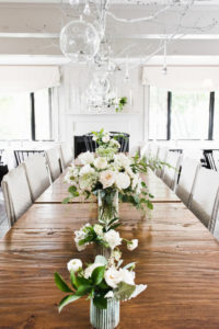 A long wooden table decorated with vases of white and green flowers, hanging glass orbs, and white branches above, set in a bright room with large windows, modern chairs, and elegant packages adorning the centerpiece.