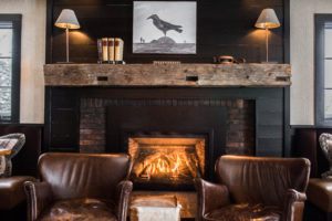 Cozy living room with a lit fireplace, two brown leather chairs, a rustic wooden mantel, books, and a painting of a black bird above the mantel—perfect for relaxing or enjoying casual fine dining by the warm glow of wall lamps.