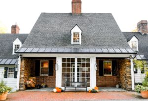 A brick house with a black shingle roof, white trim, and a small dormer window evokes the cozy charm of an Edson Hill - Stowe Vermont Inn. Pumpkins, firewood, and black lanterns decorate the inviting porch with a bench by the large window.