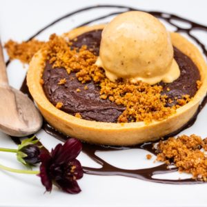 A chocolate tart topped with a scoop of vanilla ice cream, garnished with cookie crumbs, chocolate sauce, and a small dark flower, served on a white plate with a wooden spoon—an elegant addition to our Farm to Table Menu Vermont.