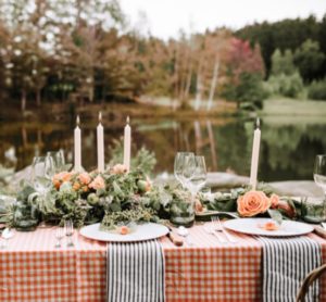 A table set for a Thanksgiving Dinner in Stowe Vermont outdoors by a lake, decorated with orange flowers, greenery, tall white candles, wine glasses, and plates on a red-and-white checkered tablecloth with striped napkins.