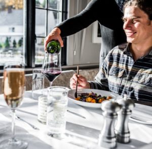 A person pours red wine into a glass at a sunlit restaurant table, where a smiling man enjoys a dish of colorful food—capturing the warmth of Thanksgiving Dinner in Stowe Vermont. Glasses and condiments are scattered across the white tablecloth.