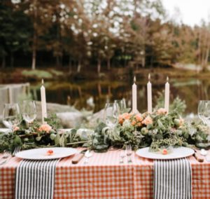 A beautifully set outdoor table for Thanksgiving Dinner in Stowe, Vermont features a red-and-white checkered tablecloth, striped napkins, floral and greenery centerpiece, candles, glassware, and plates overlooking a peaceful pond and trees.