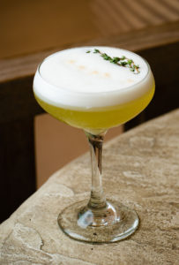 A coupe glass filled with a yellow-green cocktail topped with a layer of white foam, garnished with fresh thyme, sits on a textured stone surface—an inviting choice for your next menu.