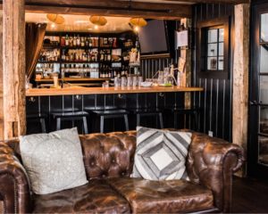 A cozy bar interior with a brown leather sofa and patterned cushions in the foreground, facing a wooden bar lined with stools. Shelves display bottles and glassware, while a visible menu and warm lighting create an inviting atmosphere.