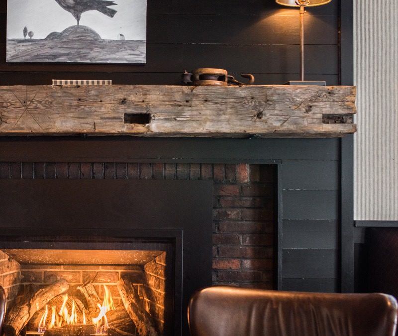 A cozy fireplace with a burning fire, rustic wooden mantel, a lamp, and a painting of a crow above creates an inviting space. Brown leather chairs are partially visible in the foreground, perfect for browsing the menu beside the dark-painted wall.