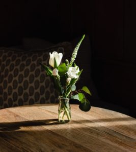 A small glass vase with white flowers and green leaves sits on a sunlit wooden table, next to a menu, with a patterned cushion and dark background behind it.