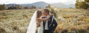 A bride and groom embrace outdoors in a sunlit field with mountains in the background, capturing the magic of weddings. The bride wears a white dress and veil, holding a bouquet, while the groom wears a dark suit.