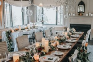 Elegant dining room set for a formal Thanksgiving Dinner in Stowe, Vermont, with long tables decorated with candles, greenery, and flowers. Natural light streams through large windows as glass orbs hang from the ceiling, creating a cozy, festive ambiance.