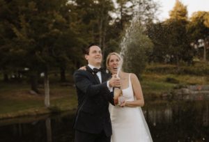 A joyful couple in formal wedding attire stands outdoors by a pond at Edson Hill - Stowe Vermont Inn, popping a bottle of champagne and laughing as the champagne sprays into the air, surrounded by lush greenery and trees.