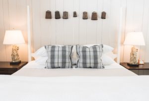 A neatly made bed with plaid pillows, white bedding, and two lamps on bedside tables. Rustic metal bells hang as wall decor above the headboard, evoking the inviting charm of Edson Hill - Stowe Vermont Inn. Clean, bright, and cozy atmosphere.