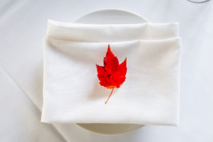 A folded white napkin with a red maple leaf on top, placed on a white plate against a white tablecloth—an inviting touch of casual fine dining elegance.