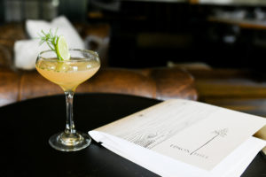 A cocktail garnished with a lime slice and rosemary sprig sits on a dark table next to an Edson Hill menu, capturing the inviting ambiance of casual fine dining against a backdrop of blurred couch and pillows.