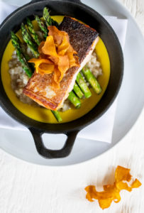 A cast iron dish with seared salmon on asparagus and creamy risotto, topped with crispy vegetable chips, sits on a white plate with a drizzle of yellow sauce—a perfect example of casual fine dining with an elegant garnish on the side.