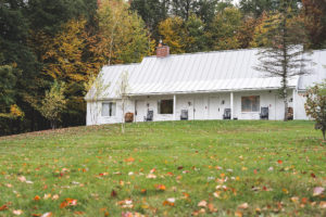 A white house with a metal roof sits on a grassy lawn blanketed in autumn leaves, surrounded by vibrant fall foliage. Four black chairs rest on the porch—perfect for relaxing during your Vermont vacation.