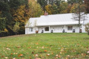 A white house with a metal roof sits on a grassy lawn, surrounded by autumn trees. Several black rocking chairs line the porch, evoking the charm of Vermont luxury hotel accommodations amid fallen leaves.