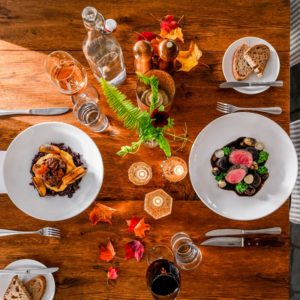 Overhead view of a wooden dining table set for two with plates of gourmet food, wine and water glasses, candles, a small green plant, and scattered autumn leaves—perfect for an intimate fine dining Stowe VT experience.
