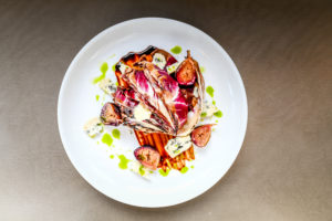 A white plate with a gourmet salad of radicchio, figs, blue cheese crumbles, and balsamic reduction, garnished with green herb oil—an elegant dish reminiscent of a Vermont Vacation—set against a neutral background.