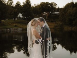Two highschool sweethearts say “I do” in Stowe as a bride and groom embrace under a sheer, flower-adorned veil by the calm lakeside, surrounded by trees at sunset—an intimate moment from Hannah & Colin’s quintessential Vermont wedding at Edson Hill.