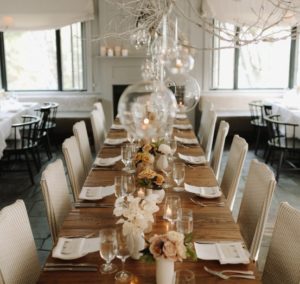 A long wooden dining table set for a formal Thanksgiving Dinner in Stowe Vermont, decorated with flowers and candles, glass globes hanging above, surrounded by cushioned chairs in a softly lit, elegant room with large windows.