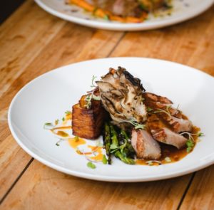A plated gourmet dish featuring sliced meat, crispy roasted potatoes, asparagus, mushrooms, and microgreens, drizzled with sauce—perfect for a Thanksgiving Dinner in Stowe Vermont—served on a white plate atop a wooden table.