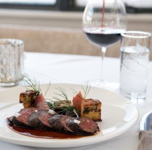 A plated gourmet meal featuring slices of seared steak with sauce, grilled potato cubes, and elegant garnishes—perfect for a refined Thanksgiving Dinner in Stowe Vermont—set on a white tablecloth beside red wine, water, and a softly glowing candle.