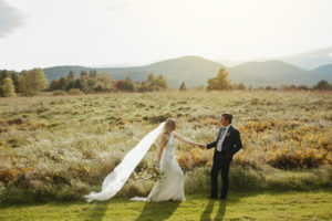 A bride in a white dress with a long veil holds hands with her groom in a suit, standing together at Edson Hill, VT, surrounded by grassy fields, mountains, and trees under soft sunlight—an Over the Moon Weddings moment.