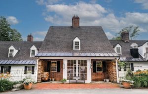 A large brick and white house with a steep gray roof, central entrance with glass doors, stacked firewood by the porch, and flowers along the pathway—evoking the timeless charm of 6 New England Vacations to Take From Westchester—under a partly cloudy sky.