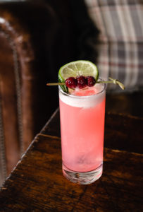 A tall glass of pink cocktail from the menu, topped with ice and garnished with a slice of lime and red berries on a skewer, sits on a wooden table with a leather chair and plaid cushion in the background.