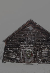 A rustic wooden barn stands in falling snow at Edson Hill - Stowe Vermont Inn, its door adorned with a holiday wreath. Snow blankets the ground, creating a wintry, festive atmosphere in this charming Vermont setting.