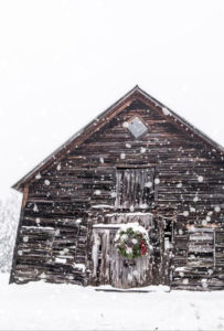 A rustic wooden barn stands in the snow at Edson Hill - Stowe Vermont Inn, adorned with a festive wreath on its door. Snowflakes gently cover the roof and ground, creating a wintry and peaceful atmosphere.